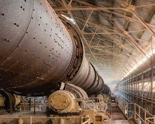 Cement plant with large white rotary kiln, cylindrical storage silos, and blue industrial buildings.