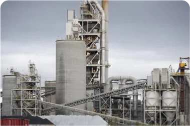 Cement plant with storage silos and processing towers surrounded by green trees.